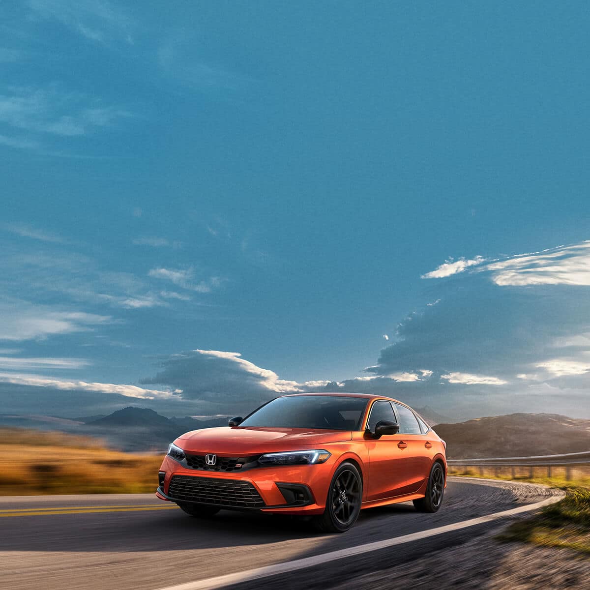 An orange car is driving on a winding road with a blue sky and clouds in the background.