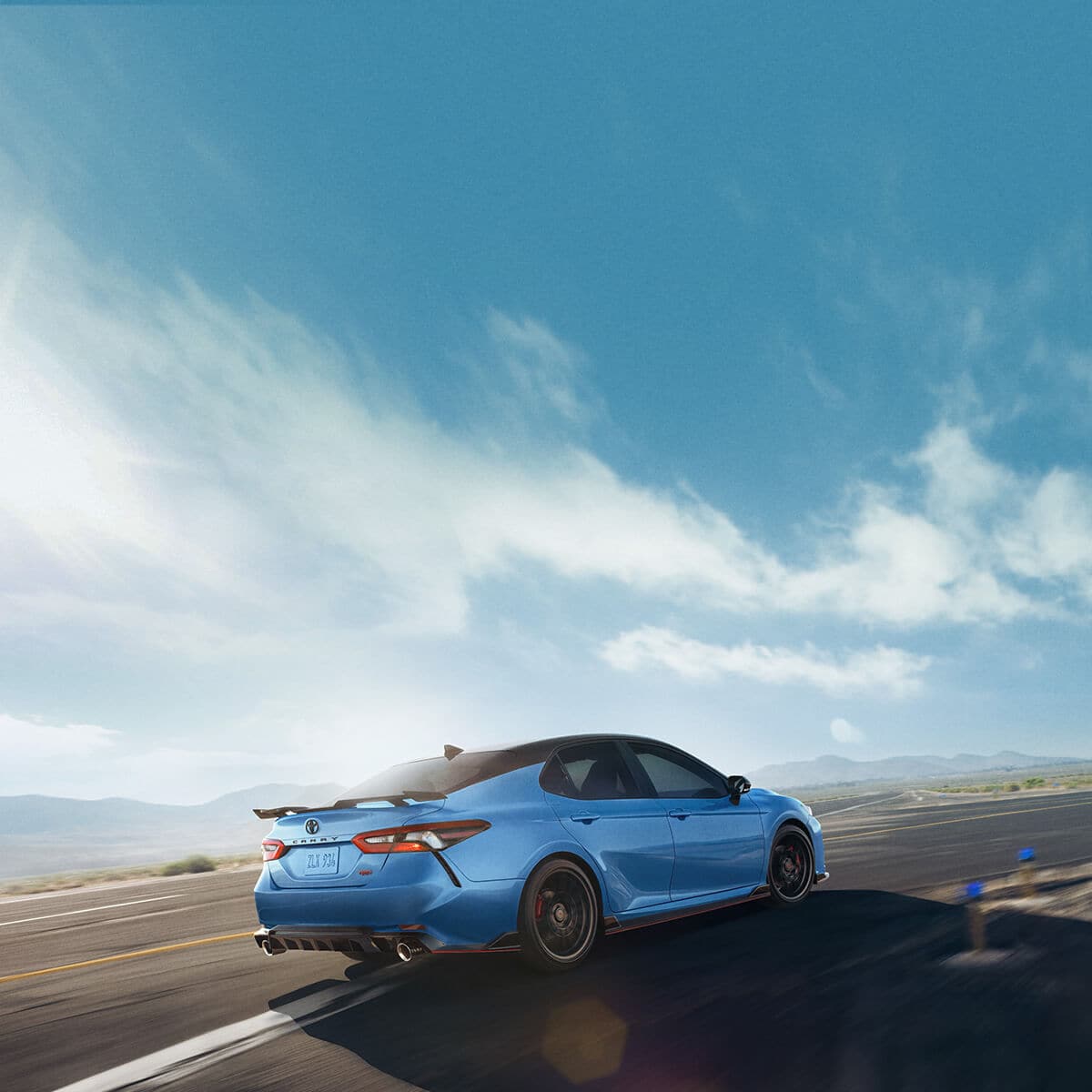 A blue sports sedan is driving on a winding road under a clear sky, with mountains in the background.
