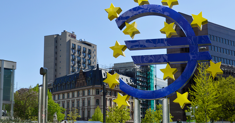 A large, blue Euro symbol sculpture stands prominently in the foreground against a clear blue sky. The sculpture is decorated with several large, yellow five-pointed stars fixed around its curved edges and central horizontal bars.  The background features an urban cityscape in Frankfurt, Germany, including:  A classic European-style building with a dark mansard roof and many windows to the left.  A tall, modern gray apartment or office building behind it.  A large, dark brown office block to the right.  Bright green trees in early spring bloom scattered between the buildings.  The lighting is bright and direct, casting soft shadows on the buildings and highlighting the reflective surface of the blue Euro sign.