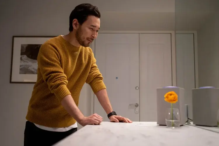 A man in a yellow sweater leans over a marble countertop, looking toward a smart speaker as if interacting with a voice assistant. The setting is a minimalist, modern room with soft lighting, reflecting the convenience of home-based voice commerce.