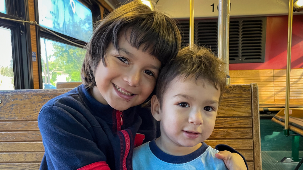 A young child hugs a younger child on a bus seat as both smile for the camera.