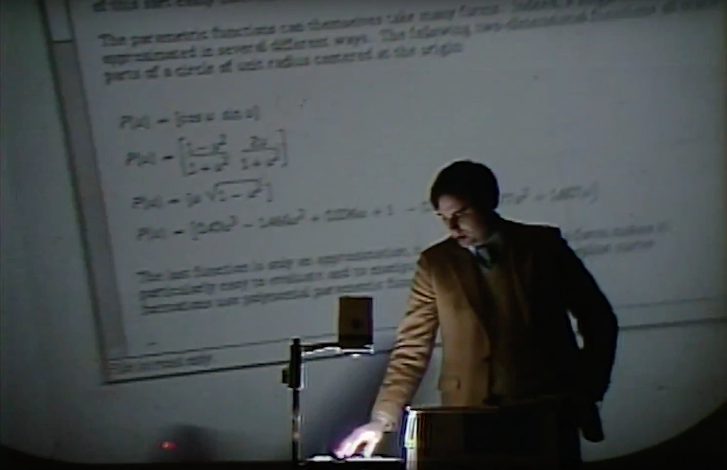 A man stands beside an overhead projector in a dimly lit classroom, text written on transparency projected on screen behind him