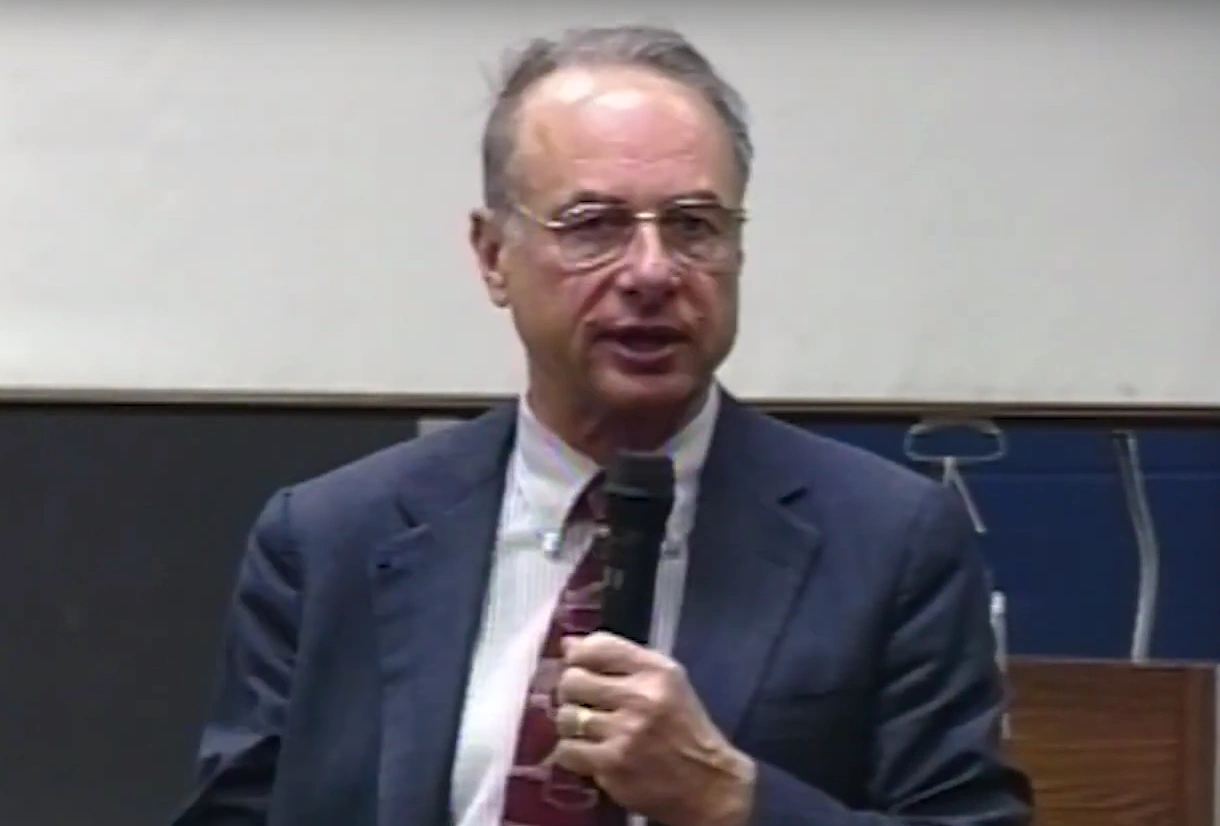 John M. Logsdon stands in front of a podium, peaking into a microphone onstage, a blank project 
