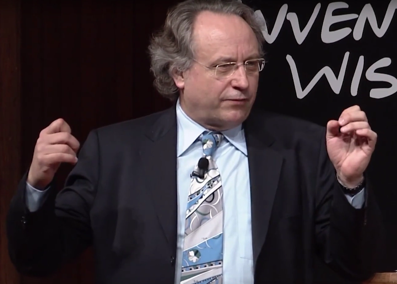 Rodney Brooks speaking onstage, a dark backdrop with white text from the MIT150 event in the background.