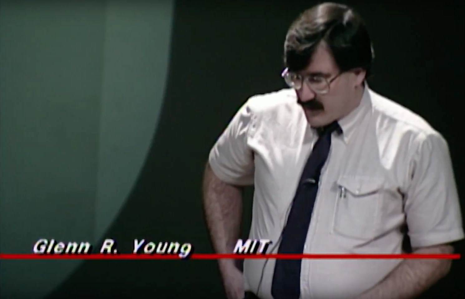 Glenn R. Young, wearing short sleeve button up shirt and tie, standing in front of green walls, looking downward