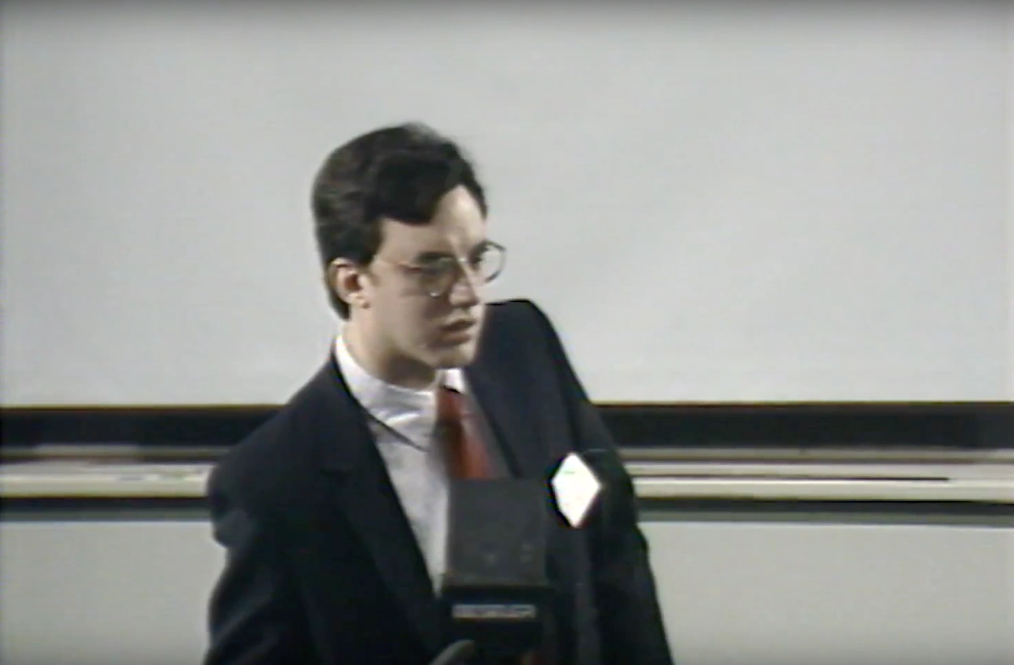 A man leans forward on podium speaking into microphone in classroom, projector screen and chalkboard in background