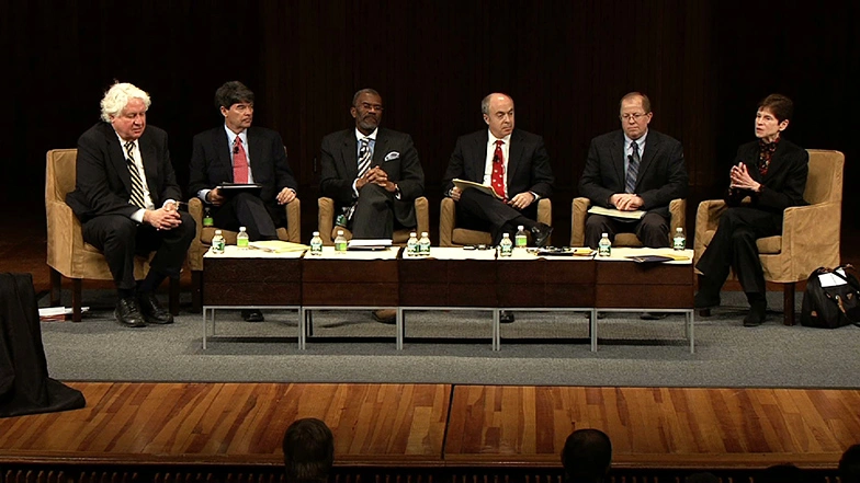 Six panelists having a discussion seated in chairs around a coffee table on stage with black background.