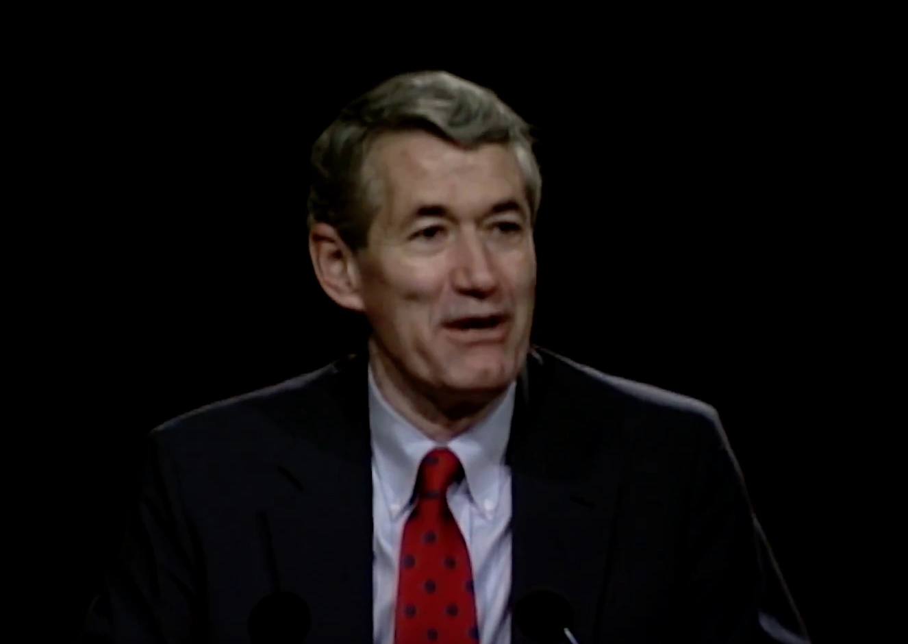Robert J. Birgeneau speaking at a podium on stage, in front of a dark background
