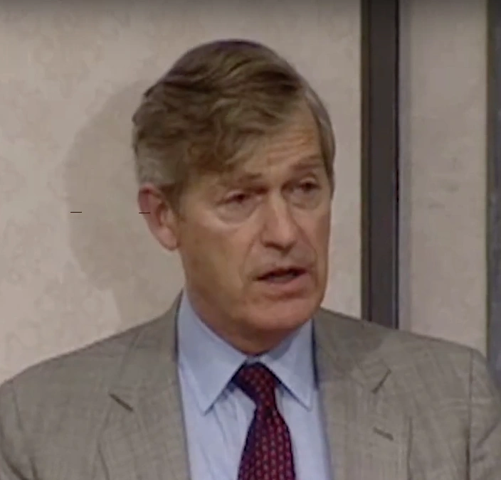 Tim Wirth speaking at a podium, cream colored wall in background