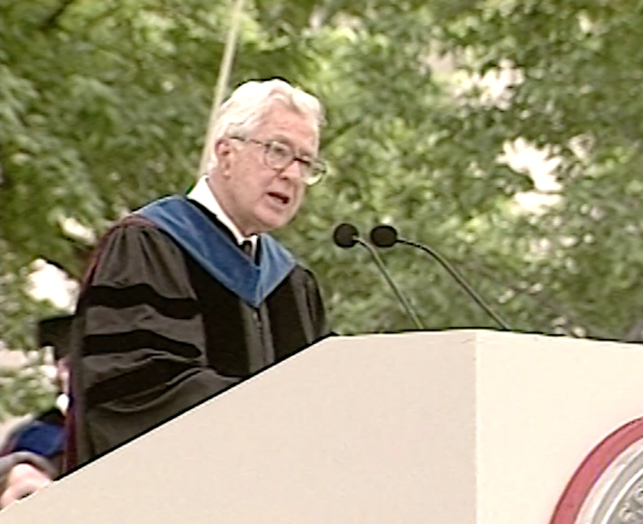 Virgilio Barco , wearing commencement attire, speaking at podium on outdoor stage, trees in background