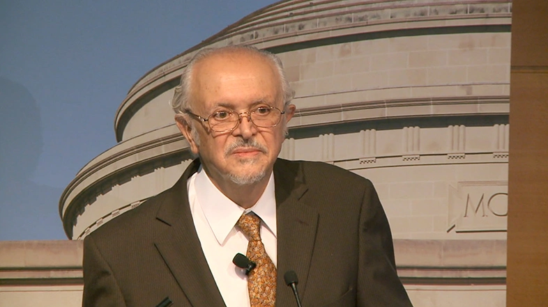 Mario Molina delivers a lecture onstage, a large image of MIT's great dome building in the background