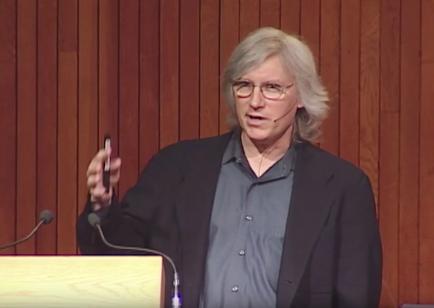 Doug Lauffenberger speaks onstage, standing behind a podium, red curtain in the background