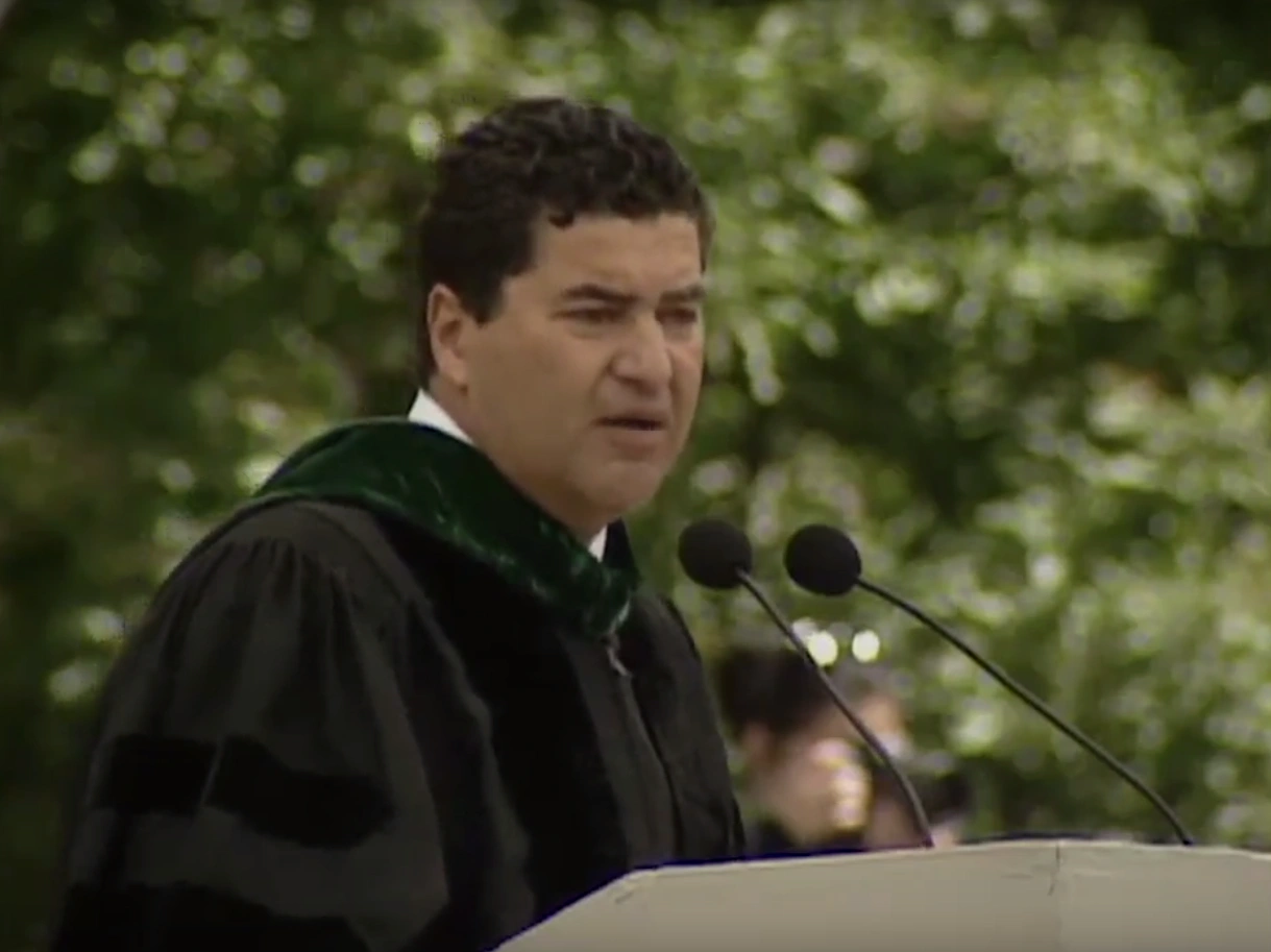 Elias A. Zerhouni, wearing commencement attire, speaking at podium on outdoor stage, trees in background