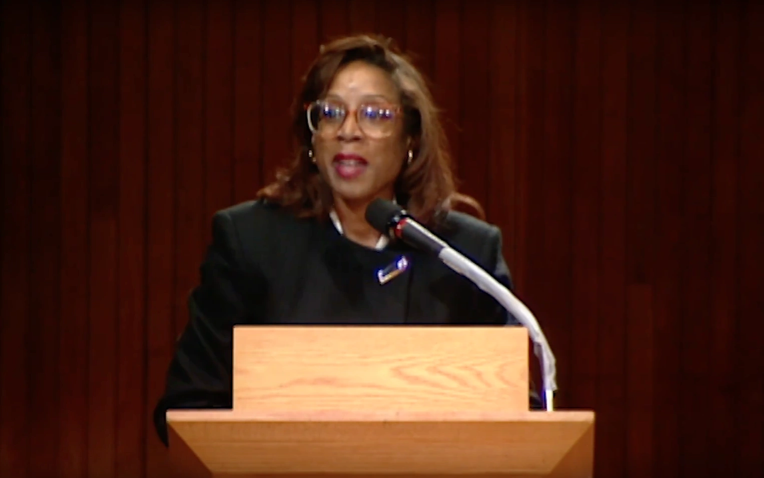Margaret Burnham speaking at a podium on stage, red curtain in background.