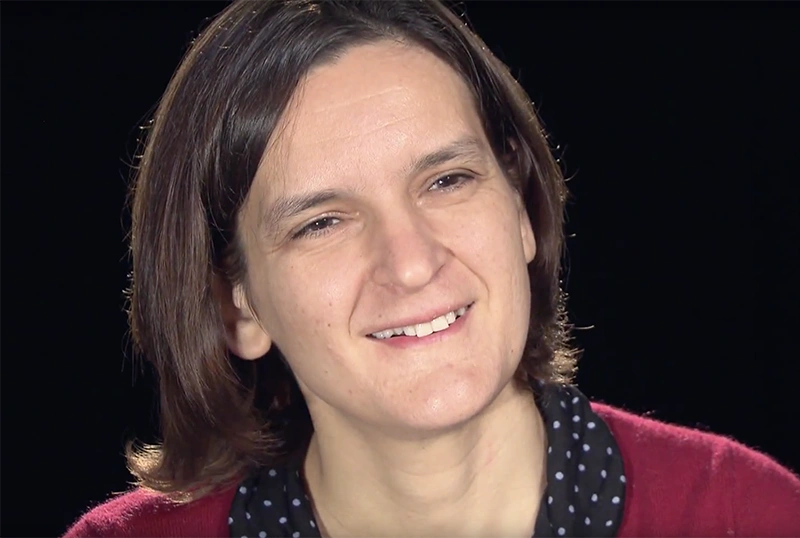 Esther Duflo seated in front of a black background for an on camera interview, smiling.