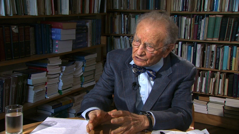 Paul Samuelson sitting at a table, large bookshelves lined with books in the background