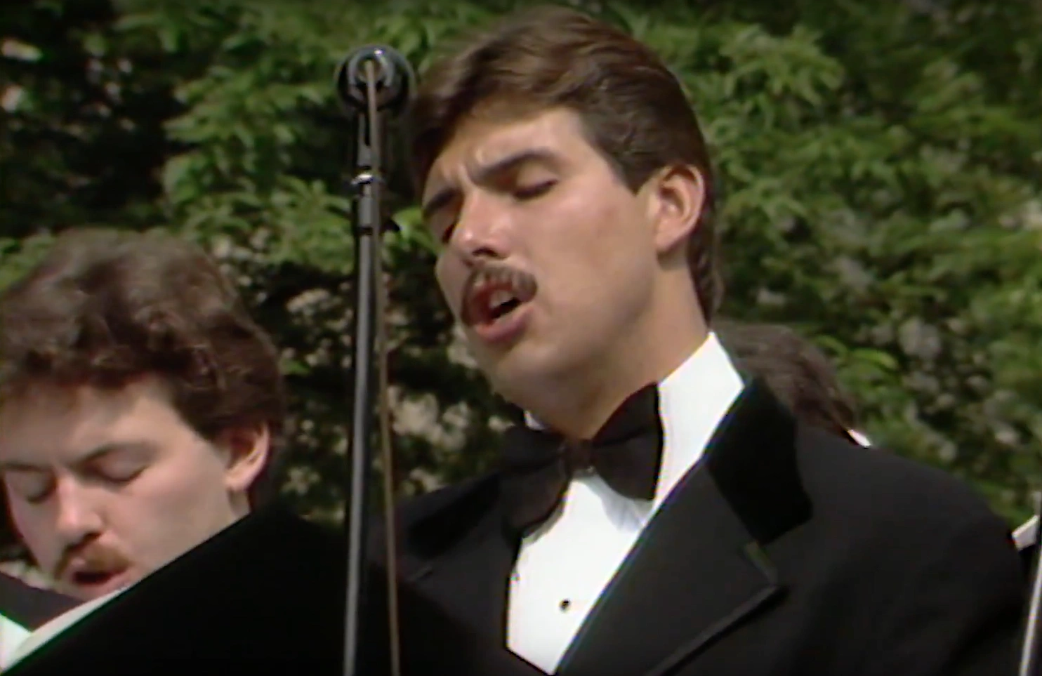 Male choir member with brown hair wearing a tuxedo, singing on outdoor stage for commencement ceremony.