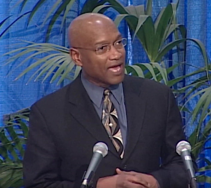 Rev. Ray Hammond speaking onstage at a podium, palm fronds and blue curtain in background