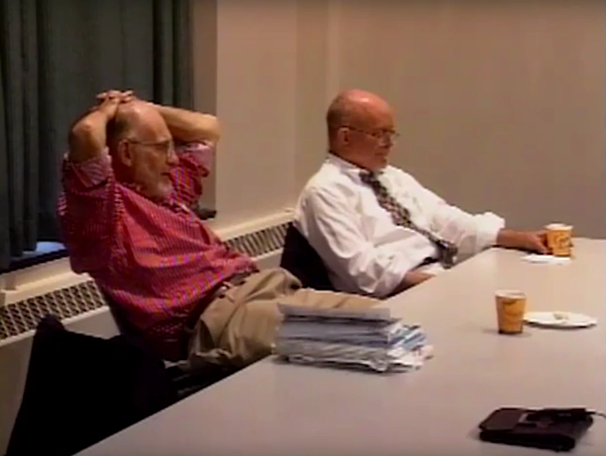 Two men sit at table in classroom for Apollo guidance computer project, one man leans back resting his head in his interlaced hands