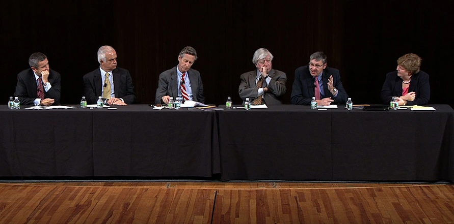 Six panelists for an economic policy panel sit at long black table on stage.