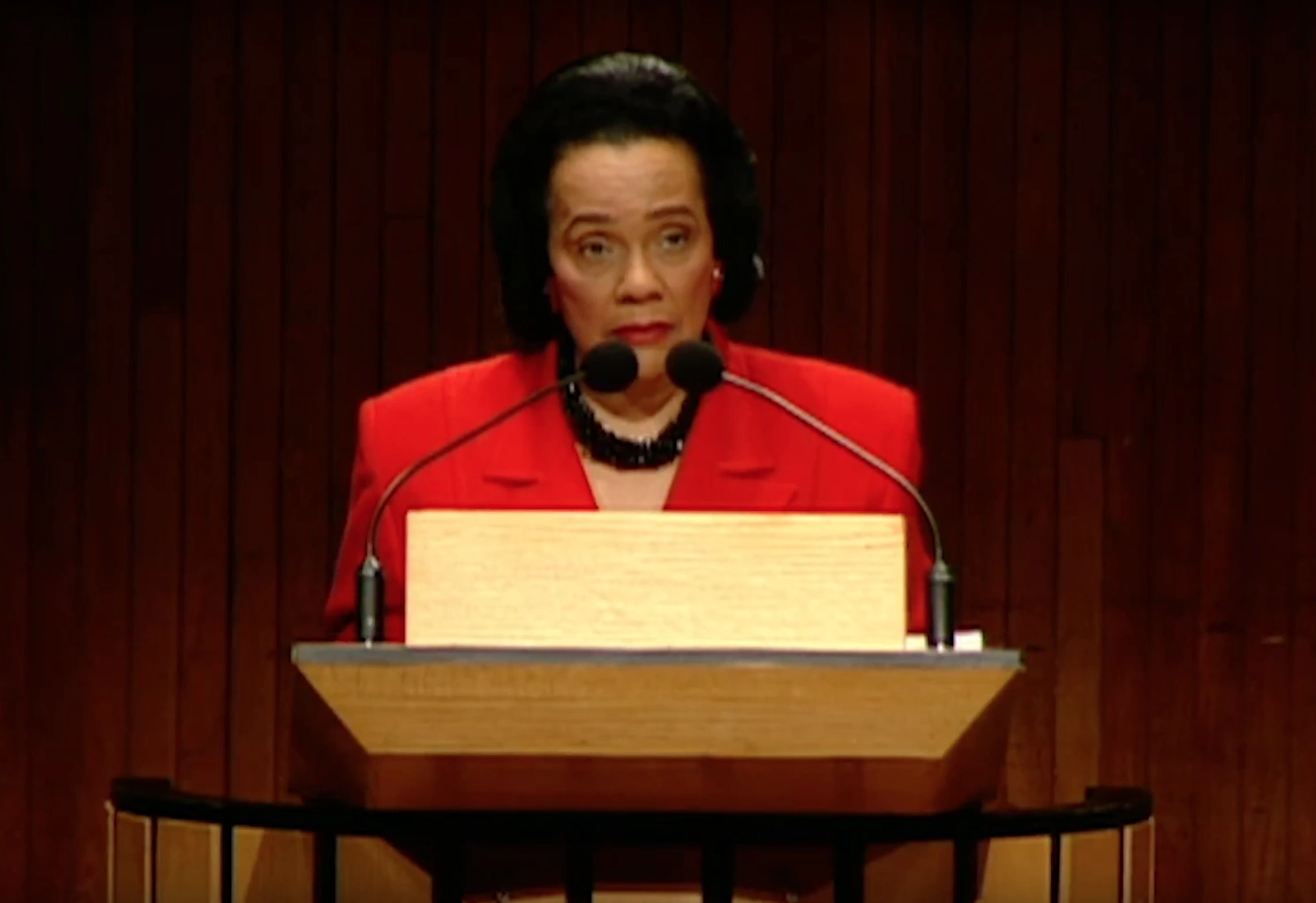 Coretta Scott King speaking at a podium on stage, red curtain in the background.