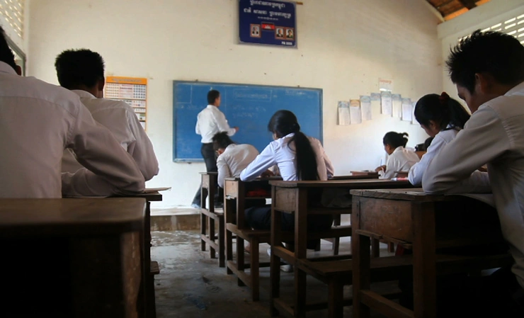A classroom with rows of students in desks looking up at their teacher at the chalkboard.