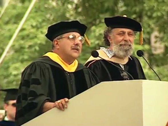 Raymond and Thomas Magliozzi, both wearing commencement attire, speaking at podium on outdoor stage, trees in background