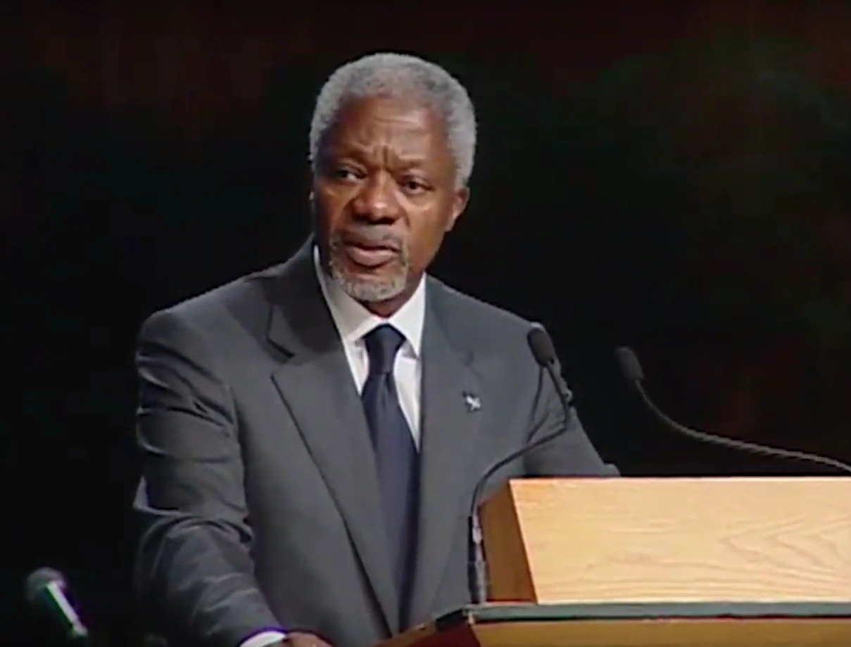 Kofi Annan speaking at a podium on stage, in front of a dark background