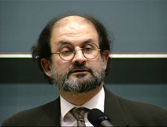 Salman Rushdie speaking at a podium in front of a chalkboard.
