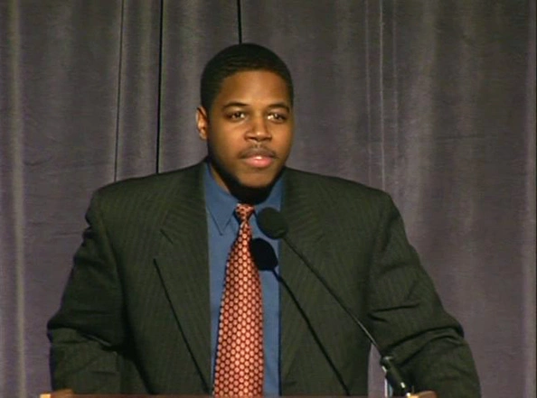 Christopher M. Jones speaking at a podium on stage, gray curtain in the background