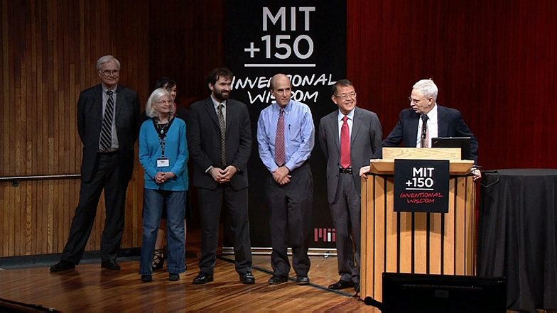 Man speaking at a podium on stage, looking at a row of five people standing on stage behind him, black backdrop