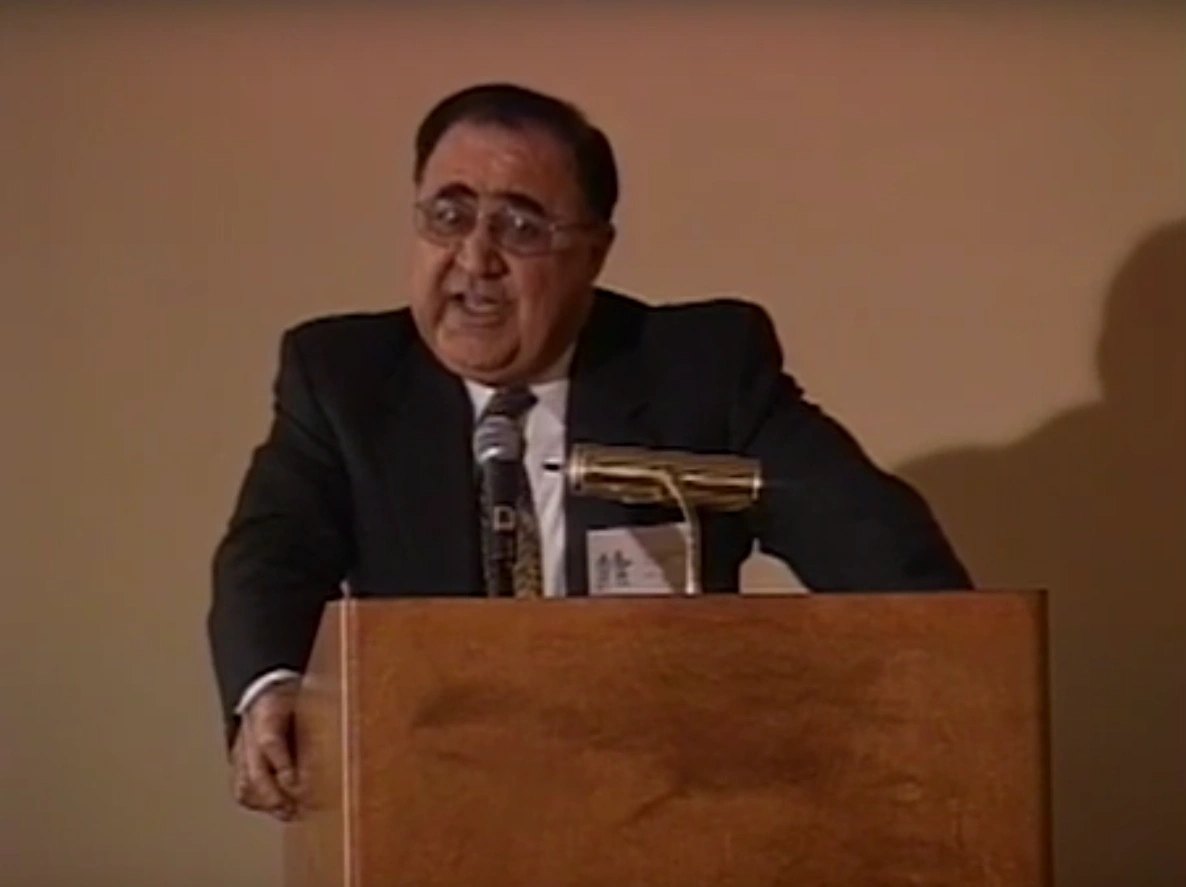 Papken Suny Der Torossian speaking at a podium on stage, in front of cream colored background.