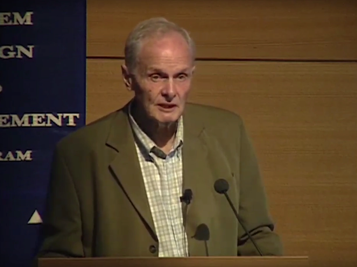 Thomas P. Hughes speaking at podium on stage, wood paneled wall in the bacground.