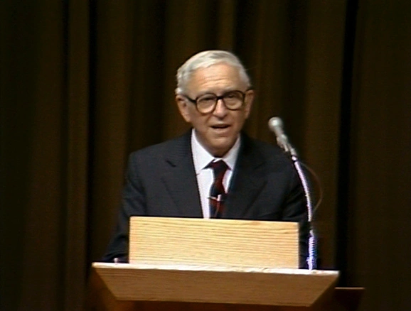 Franco Modigliani on stage speaking at podium, dark curtain in background