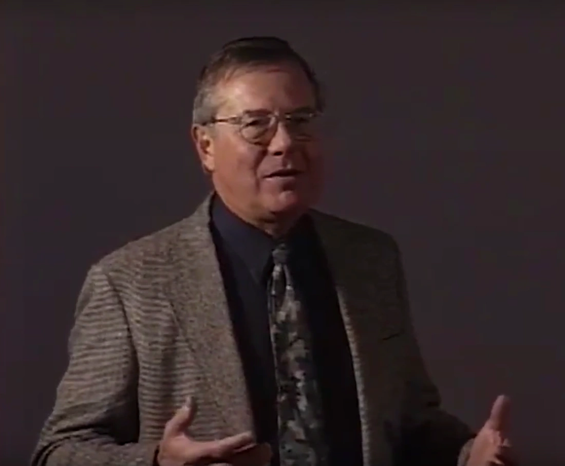 C. Forbes Dewey, Jr. speaking at a podium on stage, in front of cream colored background.