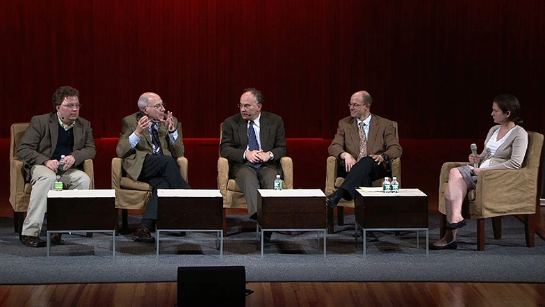 Panelists sitting side by side in upholstered chairs on stage, red curtain in background.
