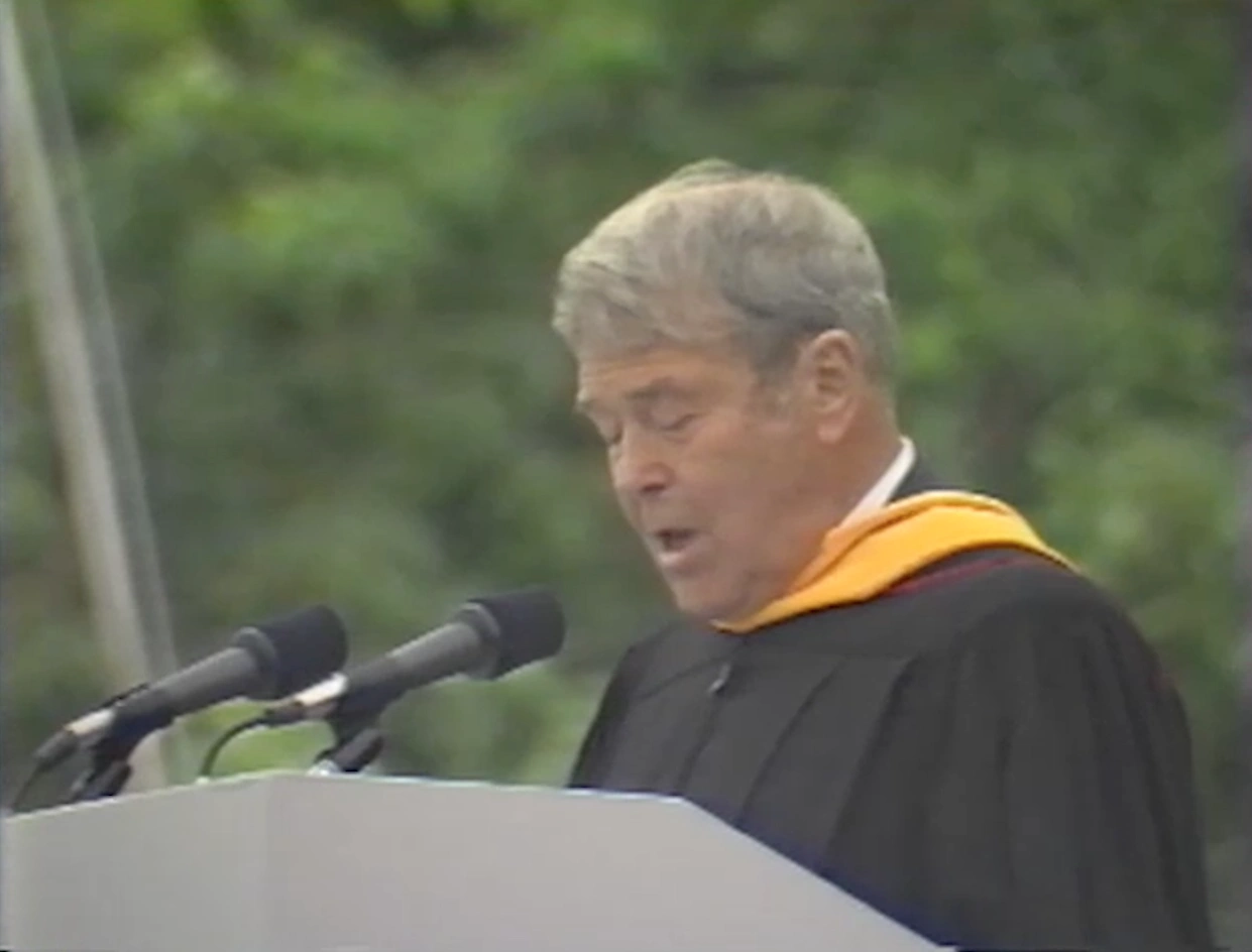 William Hewlett, wearing commencement attire, speaking at podium on outdoor stage, trees in background