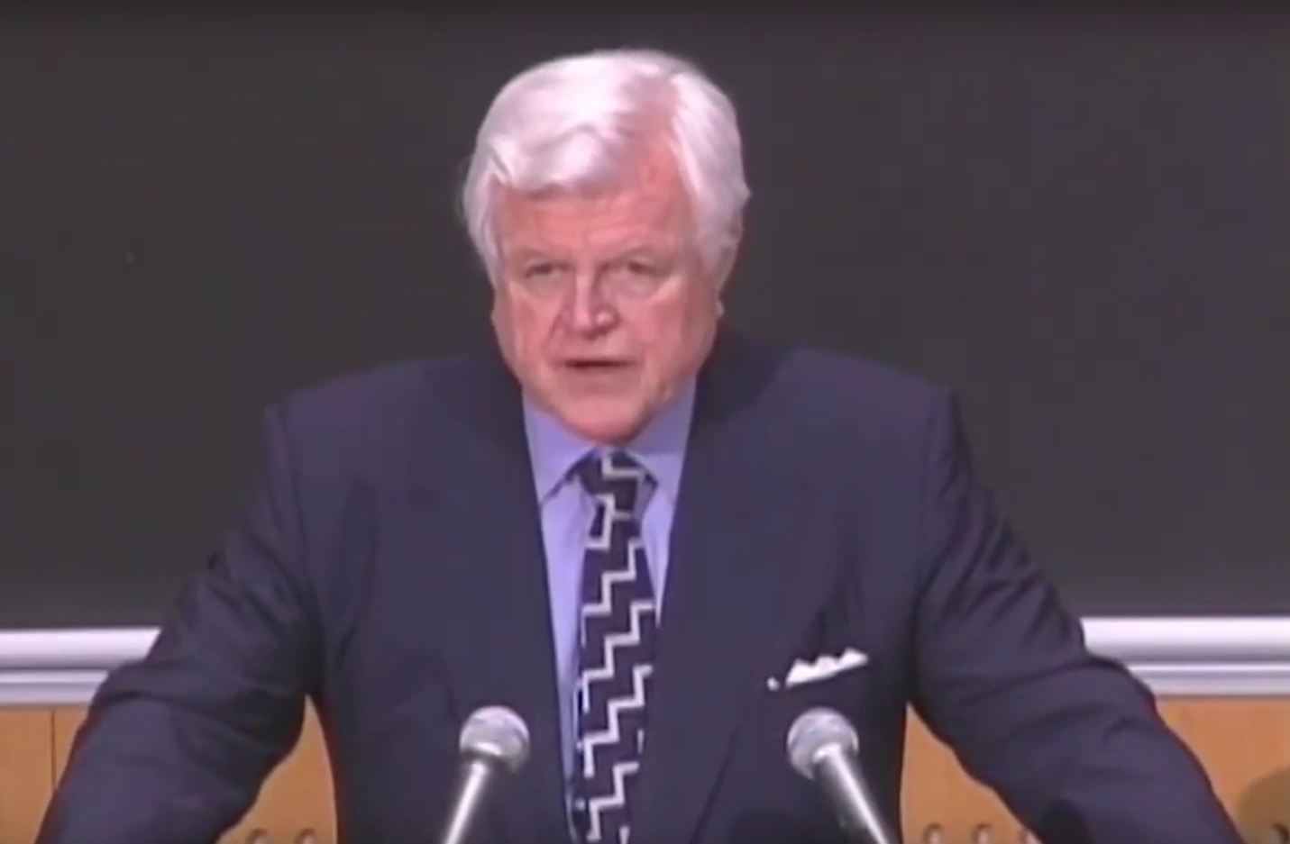 Ted Kennedy speaking into podium microphones in front of a chalkboard