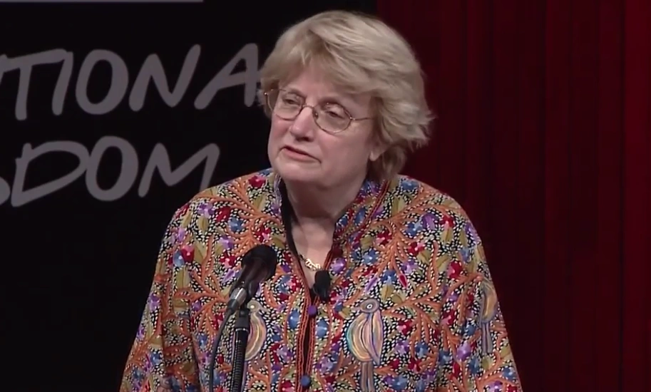 Susan Hockfield speaking at a podium on stage, a dark backdrop with white text from the MIT150 event written on it in the background.