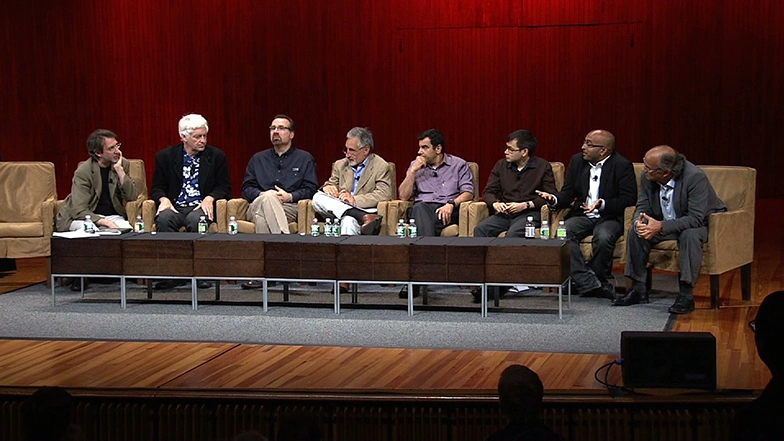 A nine person panel sit side by side in upholstered chairs on stage, red curtain in background