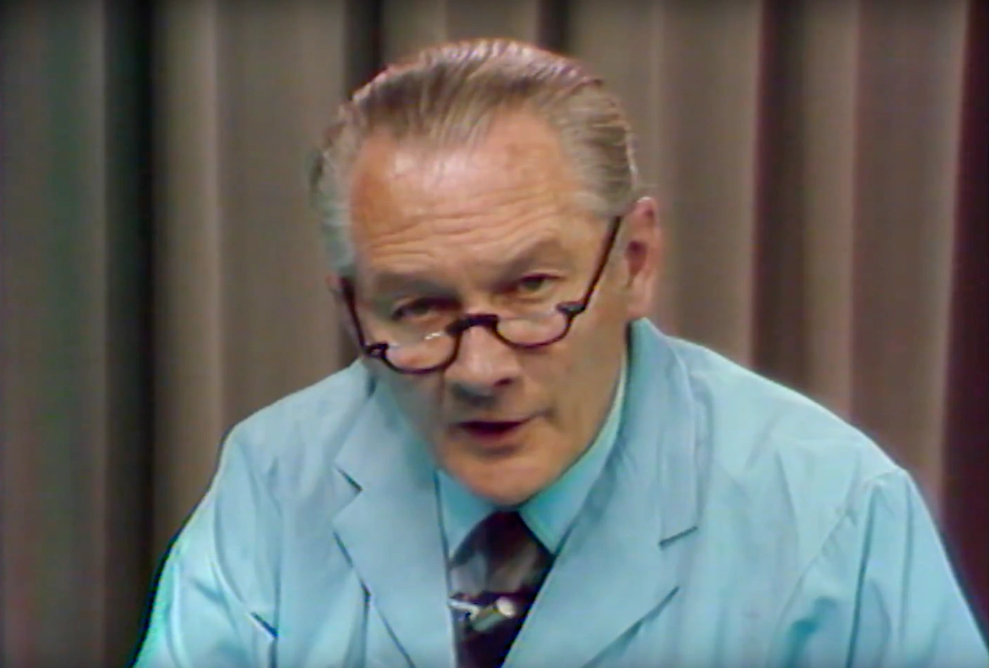 Walle J. H. Nauta, wearing blue lab coat, stands on stage, cream curtain in the background.