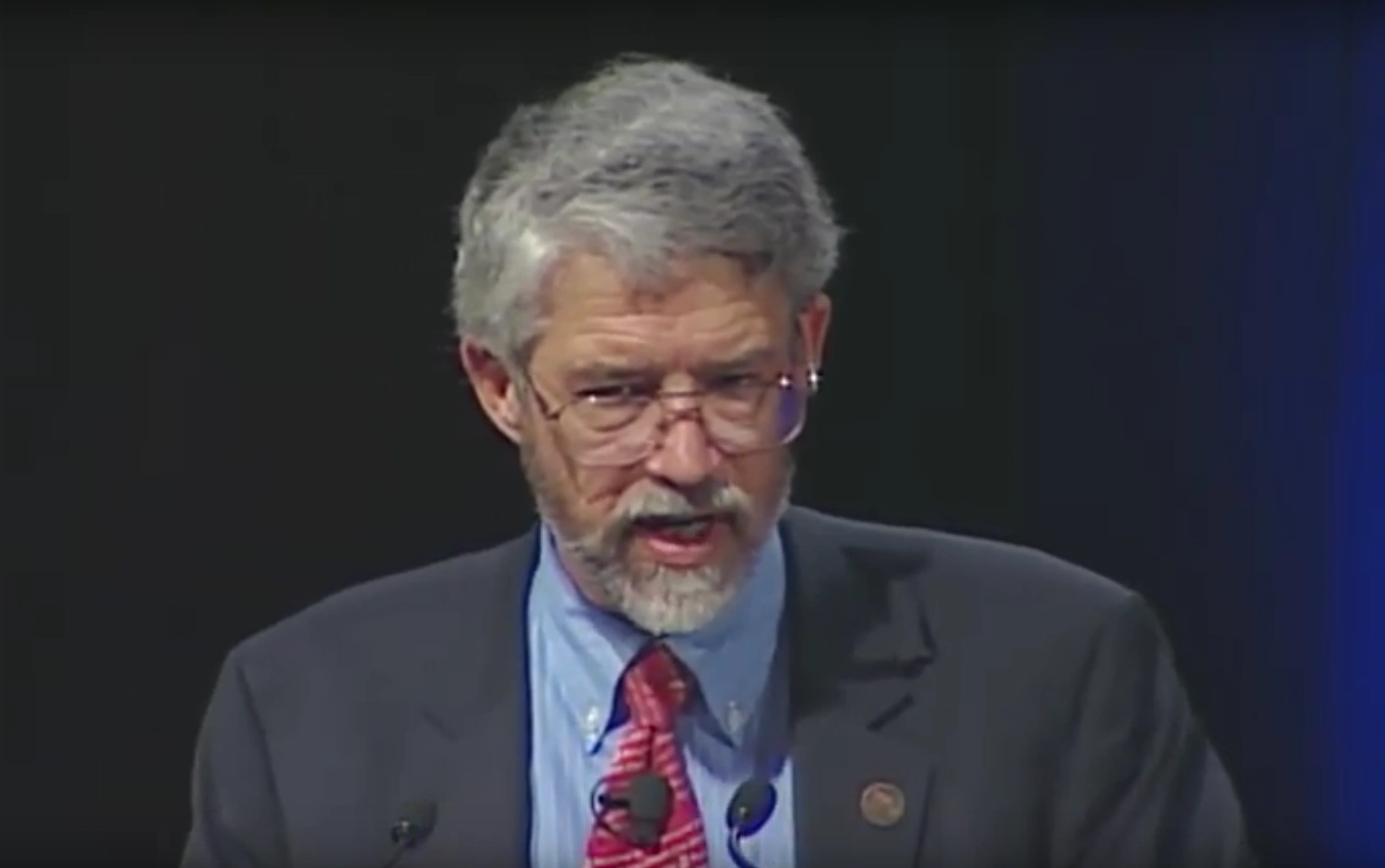 John P. Holdren speaking at podium on stage, blue curtain in background