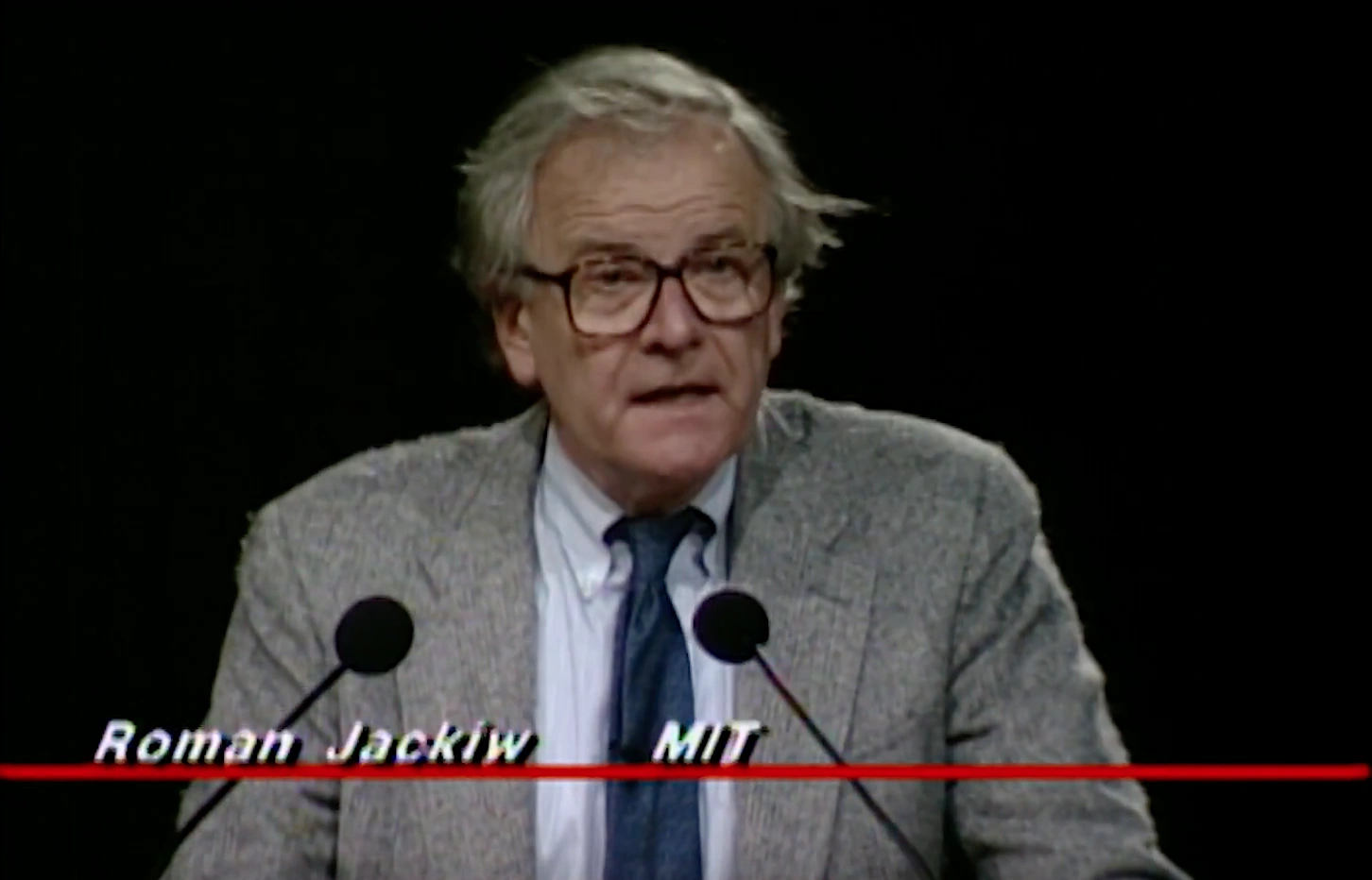 Roman Jackiw speaking at a podium on stage, in front of a dark background