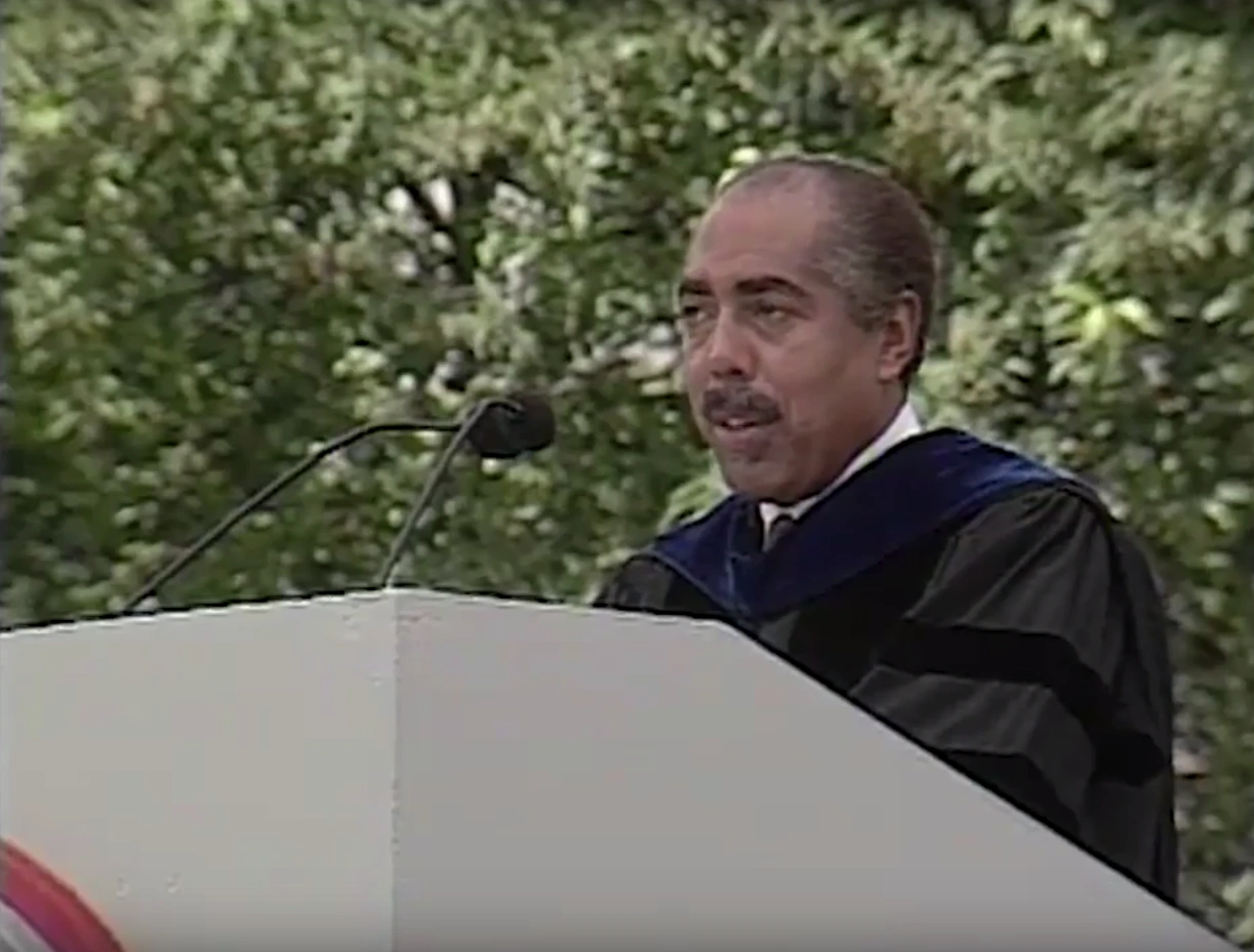 Walter Massey, wearing commencement attire, speaking at podium on outdoor stage, trees in background