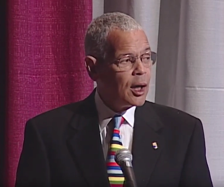 Julian Bond speaking at a podium with red curtain and columns in the background.