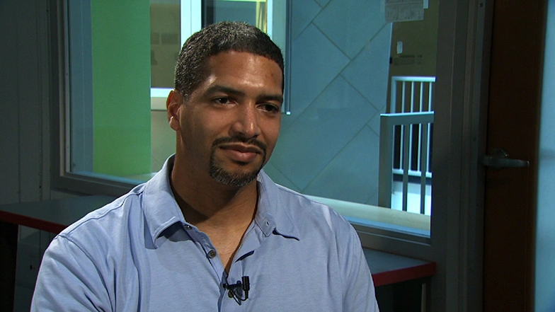 James D. McLurkin smiling during an on camera interview, seated in front of a window looking into a hallway with railings,.