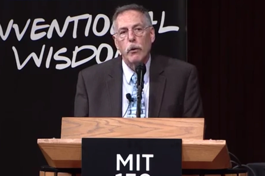 Peter Diamond speaks at a podium onstage, standing in front of behind a dark backdrop.