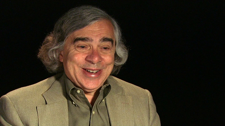 Ernest Moniz seated in front of a black background for an on camera interview, smiling.