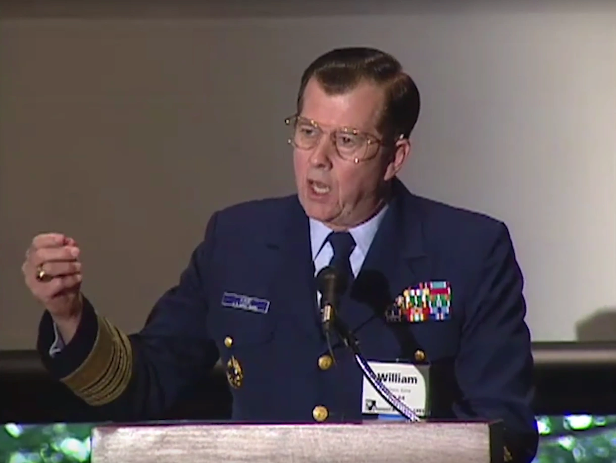 Admiral Bill Kime, wearing Marine Corps dress blues, speaks at a podium, white projector screen in background