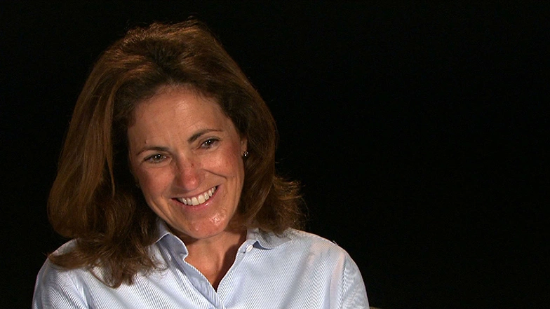 Cynthia Barnhart seated in front of a black background for an on camera interview, smiling.